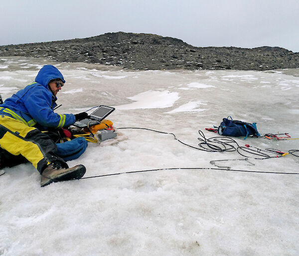 Scientist sitting on ice-covered rocks with a laptop to monitor seismic tests.