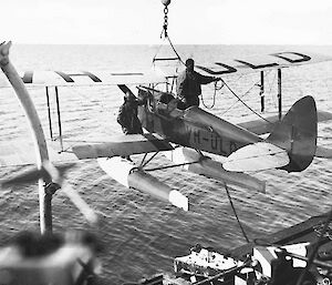 A black and white image of a biplane suspended from the side of a ship.