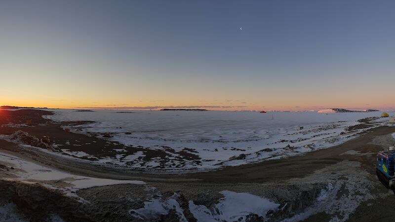 A panoramic view taken from Davis station of the sun low over Prydz Bay, with the Aurora Australis and a hagglund in view.