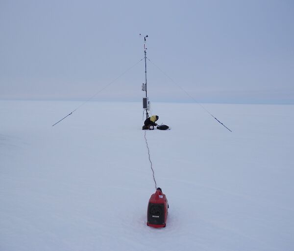 Expeditioner leans over laptop in snow underneath a weather tower (AWS)