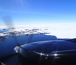Picture from the window inside of an aircraft looking at the rotating wing propeller and a glacier on the ground
