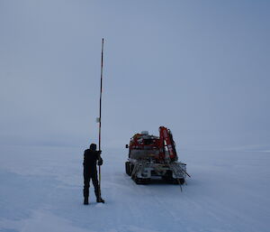 Expeditioner holds long cane into ground, surrounded by snow and ice, with vehicle just behind and to right