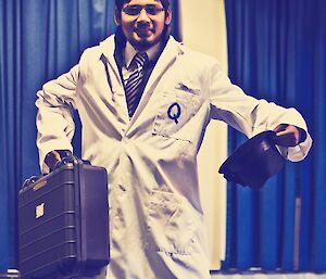 Abrar wearing a lab coat with a Q on the breast pocket. Holding a brief case and a hat
