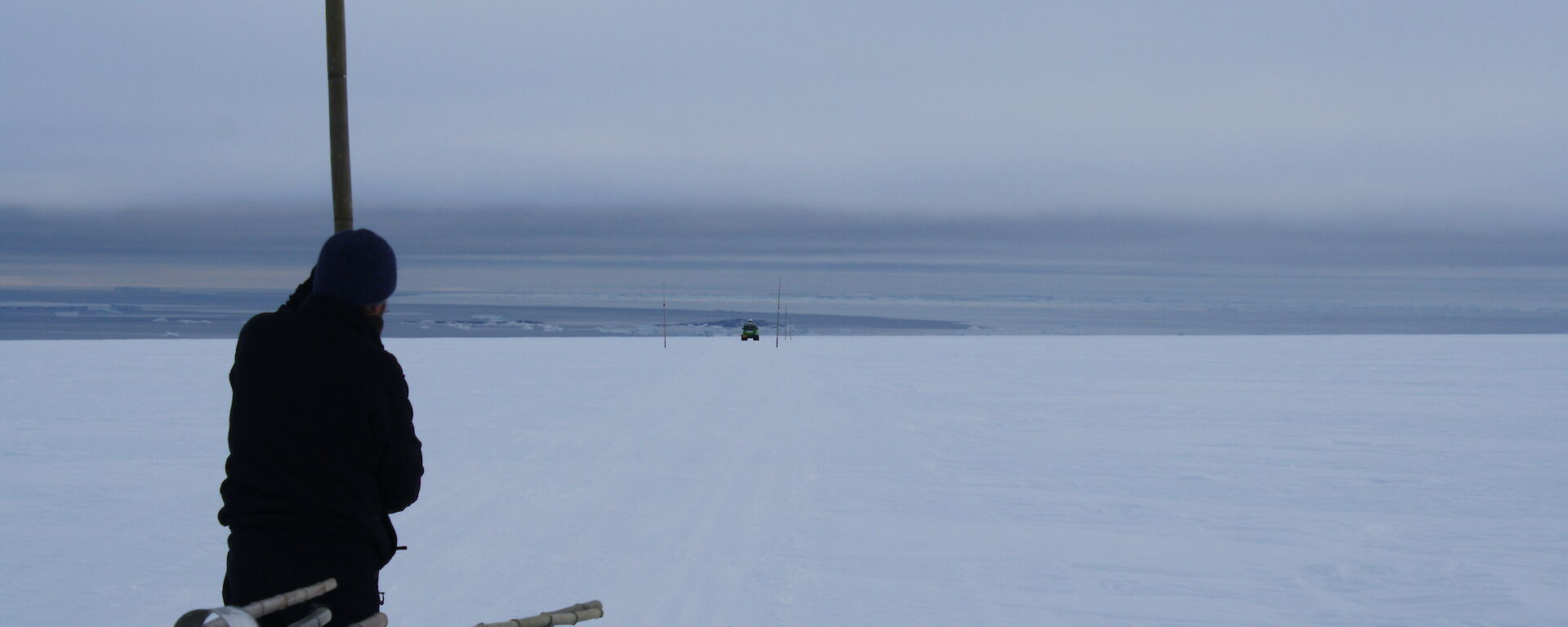 Cloudy and white flat snowy terrain, with an expeditioner standing with a cane in the foreground