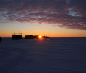 Sunrise over the skiway, a flat snow terrain with a number of vehicles and a hut