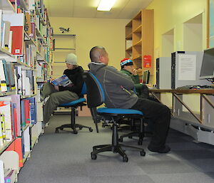 Chad, Ben and Mike relaxing in a library, Ben skimming through a book, Chad on the computer and Mike relaxing on the chair listening to the radio