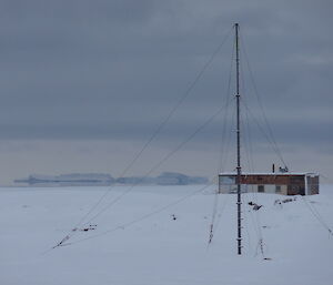 Snow covered terrain with radio antenna in the foreground, Wilkes hut in the mid ground and a large Ice berg in the background