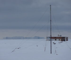 Snow covered terrain with radio antenna in the foreground, Wilkes hut in the mid ground and a large Ice berg in the background