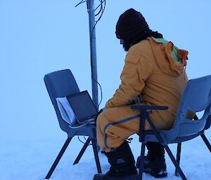 Leon sitting under the AWS in snow flat terrain, connected a laptop to the AWS