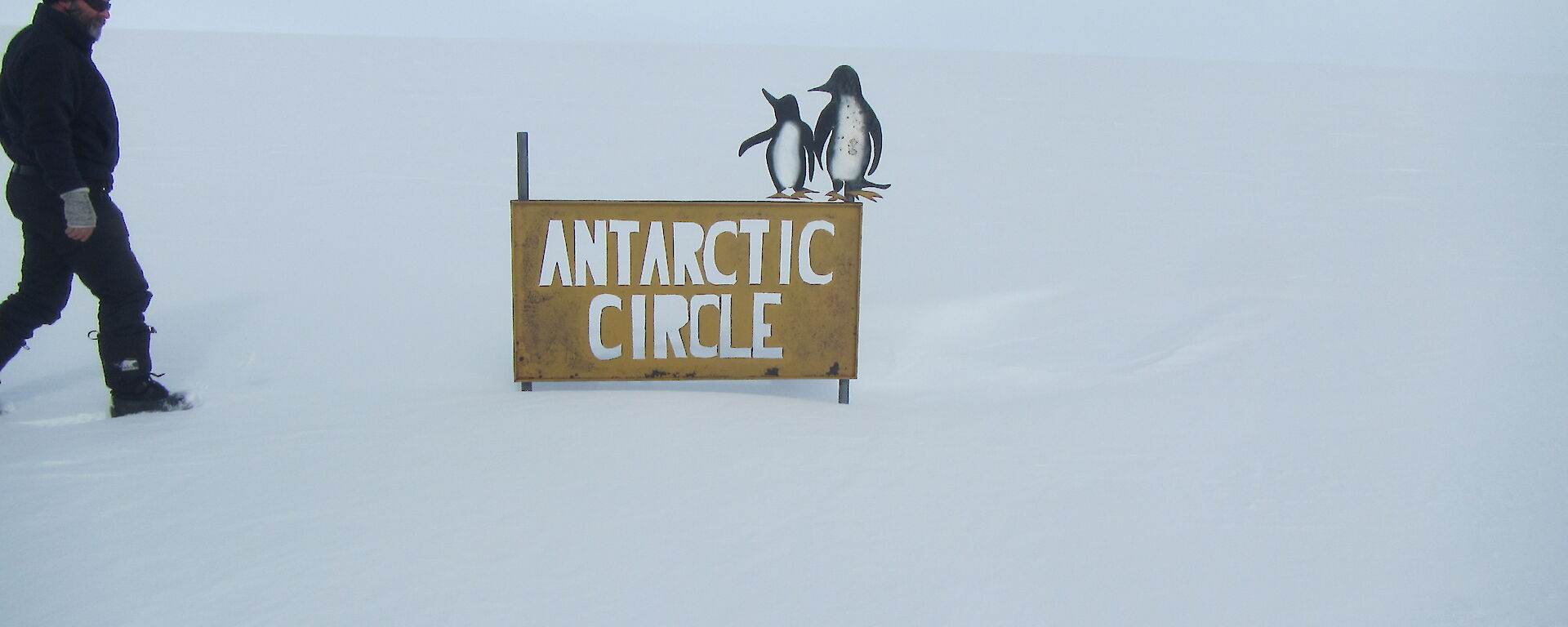Arctic Circle sign in a flat snowy terrain with Gavin walking towards it