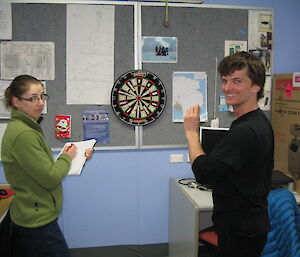 One woman holds a clipboard while the other throws darts at a dartboard
