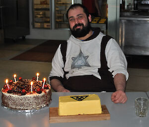 A man with a beard sits with two birthday cakes in front of him, one with lit candles