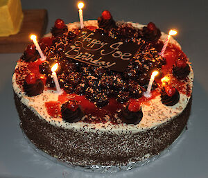 Close up of a black forest birthday cake that says ‘Happy Birthday Joe’ and is covered with lit candles