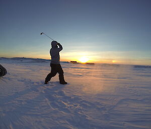 The sun is low in the sky as an expeditioner is seen just after hitting a golf ball, his club raised high, he is surrounded by ice