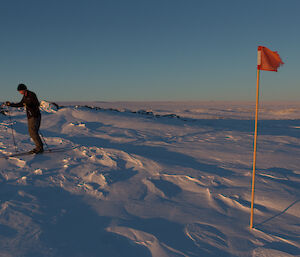 An expeditioner on skis, surrounded by snow and ice, with a flag pole to the right of the image