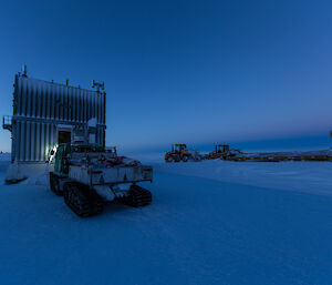 Outside at Wilkins aerodrome, near Casey station, Antarctica, shows a small building and three tracked work vehicles on the flat ice