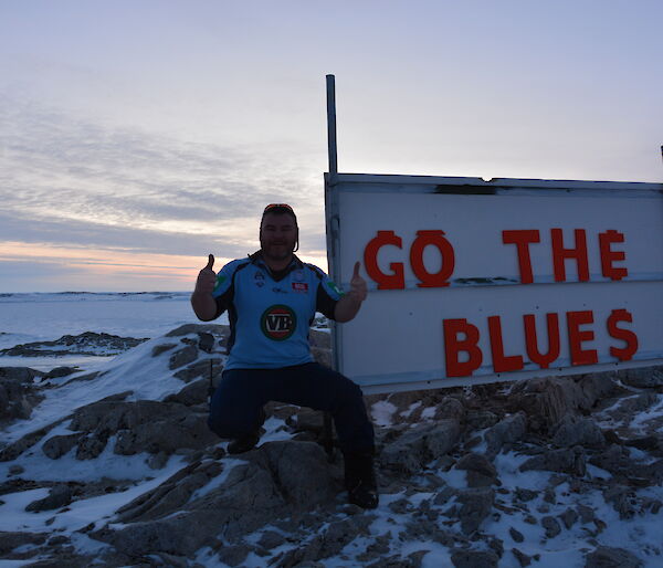 Rob Bennett sitting next to, and giving a thumbs up by, the old wooden birthday sign with his ‘Go the Blues’ message at Casey