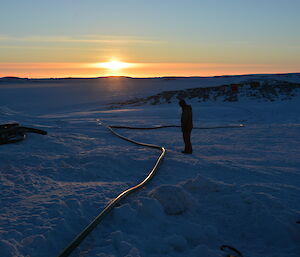 An expeditioner stands next to a hose in the snow with sun low on the horizon