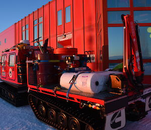 A large red tracked vehicle sits outside a red building in Antarctica, the tray holding supplies for refuelling emergency response such as fuel drums and machinery