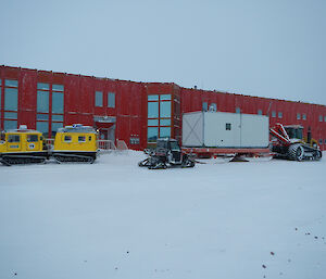 Two large vehicles outside the large red shed living quarters, the larger towing a small shed
