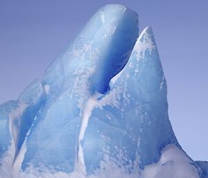 Closeup photo of a blue iceberg extending into the blue sky, with cracks and marks evident — it almost looks like a large whale has frozen while breaching with its mouth open and pointed to the sky