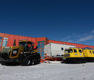 The traverse vehicles (a tractor and a Hägglunds) with sleds parked in front of the Casey station red shed on a sunny clear day.