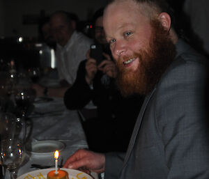A happy expeditioner at Casey station with a beard and wearing a blazer has his hands either side of his birthday dessert featuring one lit candle