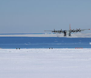A C-130 Hercules is about to touch down on the Casey skiway, with a view of sea ice and icebergs behind it.