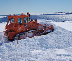 An expeditioner pushes snow away in a bulldozer, to a backdrop of the blue waters of Newcombe bay.