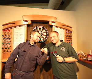 Two men posing in front of dart board