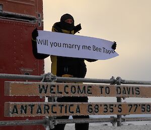 Mystery person holds up a “Will you marry me?” sign for a friend