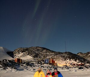 Four expeditioners stand in front of Platcha Hut beneath an Aurora