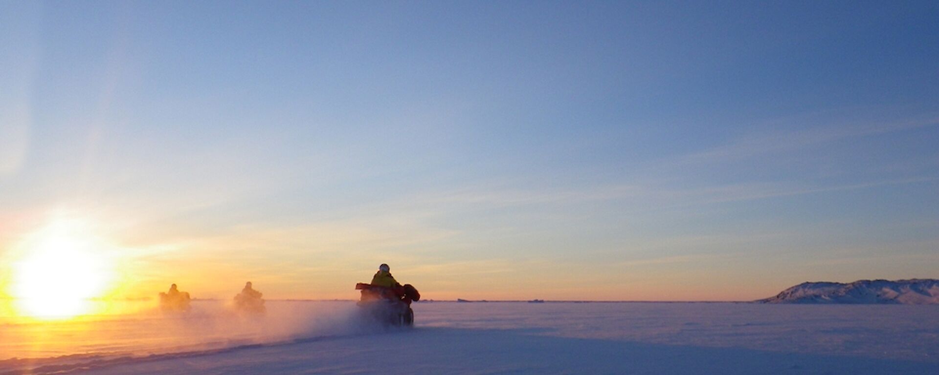 Expeditioners on three quads riding on the sea ice into the rising sun