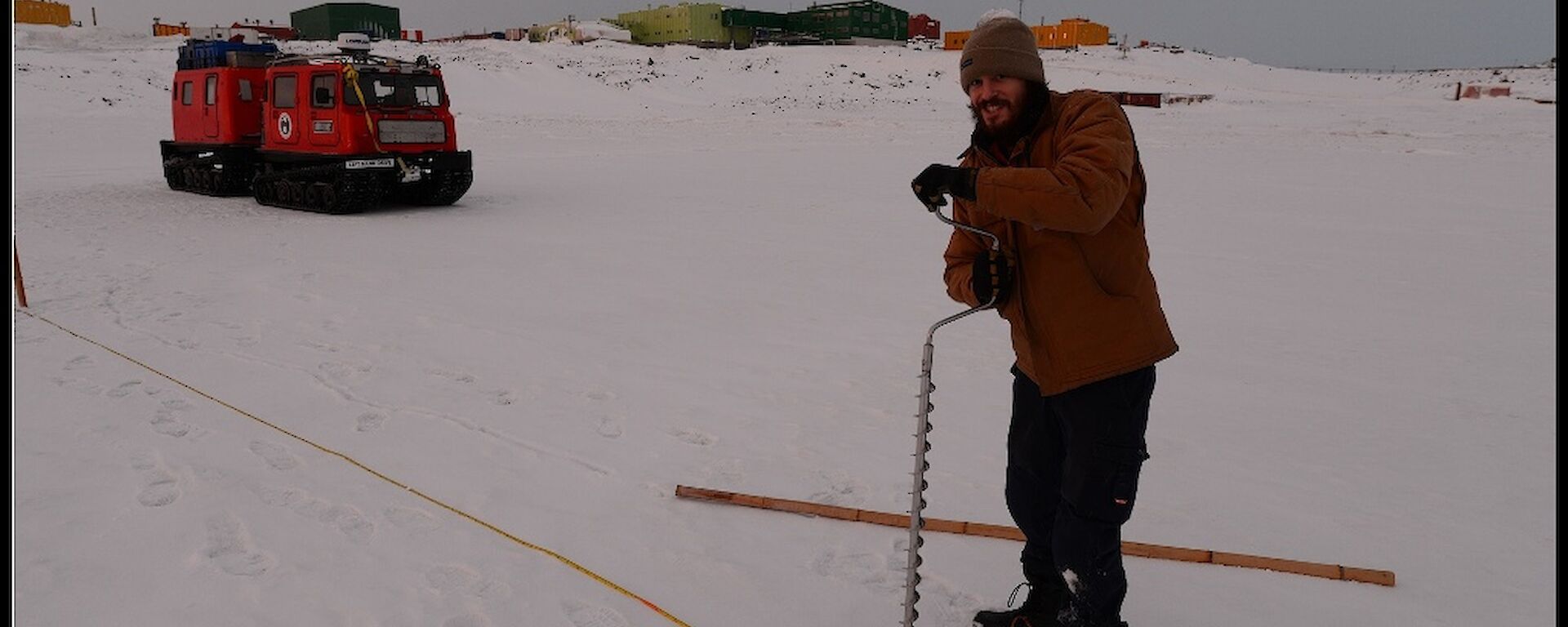 Expeditioner drilling hole in the sea ice with Hägglunds vehicle and station in the background