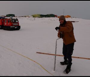 Expeditioner drilling hole in the sea ice with Hägglunds vehicle and station in the background
