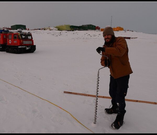Expeditioner drilling hole in the sea ice with Hägglunds vehicle and station in the background