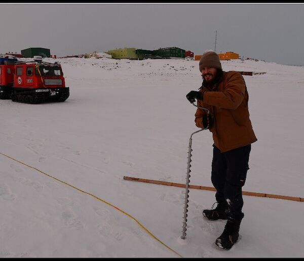 Expeditioner drilling hole in the sea ice with Hägglunds vehicle and station in the background