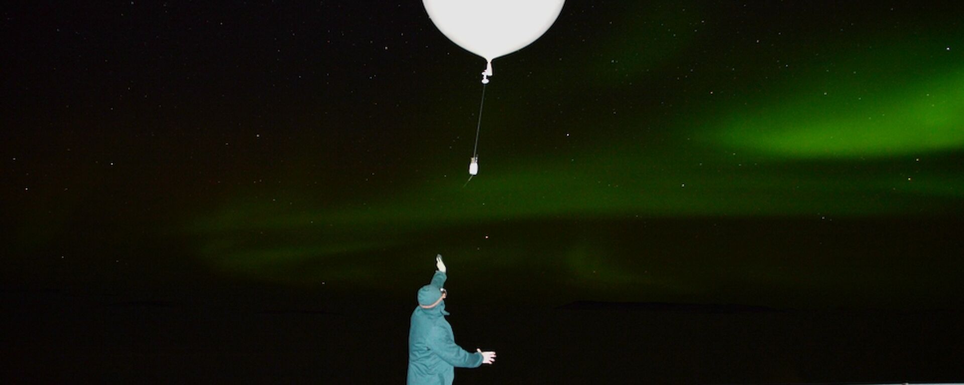 Observer wearing protective attire releasing an 800gm balloon with radiosonde, green Aurora Australis in the background