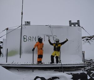 Two expeditioners stand in front of the word “Bandits” written in the snow on the side of a hut