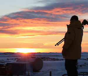 Expeditioner with camera tripod over shoulder walks in front of rising sun