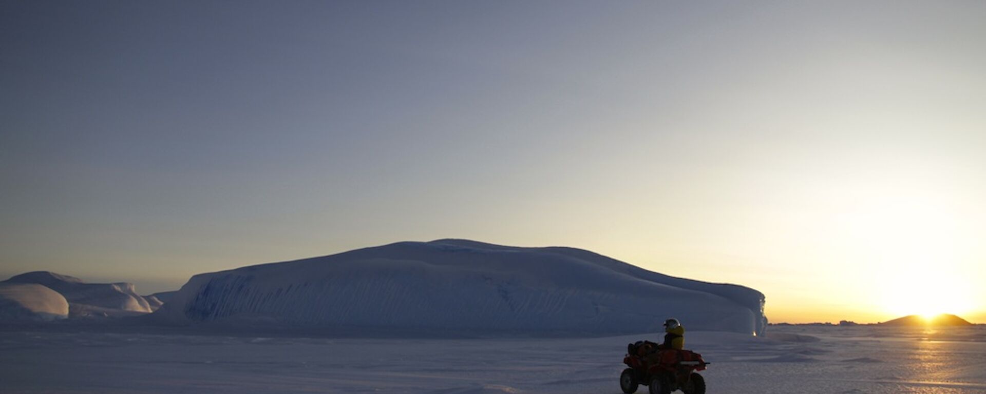 A large ice berg stuck in the fast ice in the distance with a quad bike and rider in the foreground