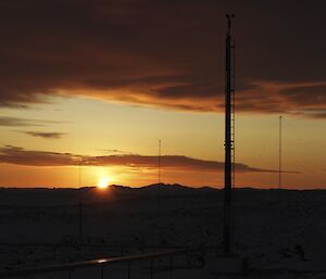 The sun rises above the horizon in clear skies with the weather station mast, antenna masts and cloud cover in the foreground