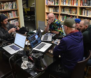 A group of expeditioners crowd together in the library to discuss the finer points of editing the movie. The table in front of them is littered with computers, cameras, radios and food