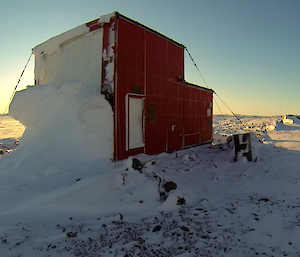 Picture of the red Band Hut, a large bit of snow blizz peeling off the left side of the building with the bright sun lighting up the frozen sea ice behind it