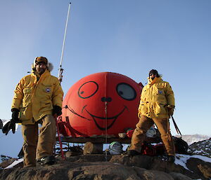 Two expeditioners standing in front of the melon hut which has a big smiley face painted on it