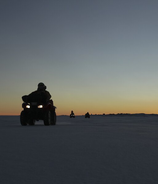 Expeditioners on the sea ice on quads with sunrise and Davis Station in the background