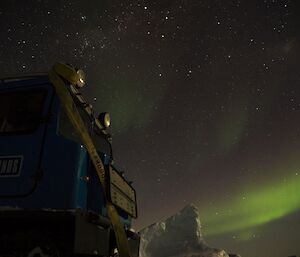 The blue Hägglunds vehicle parked on the sea ice with an ice berg and green aurora in the distance