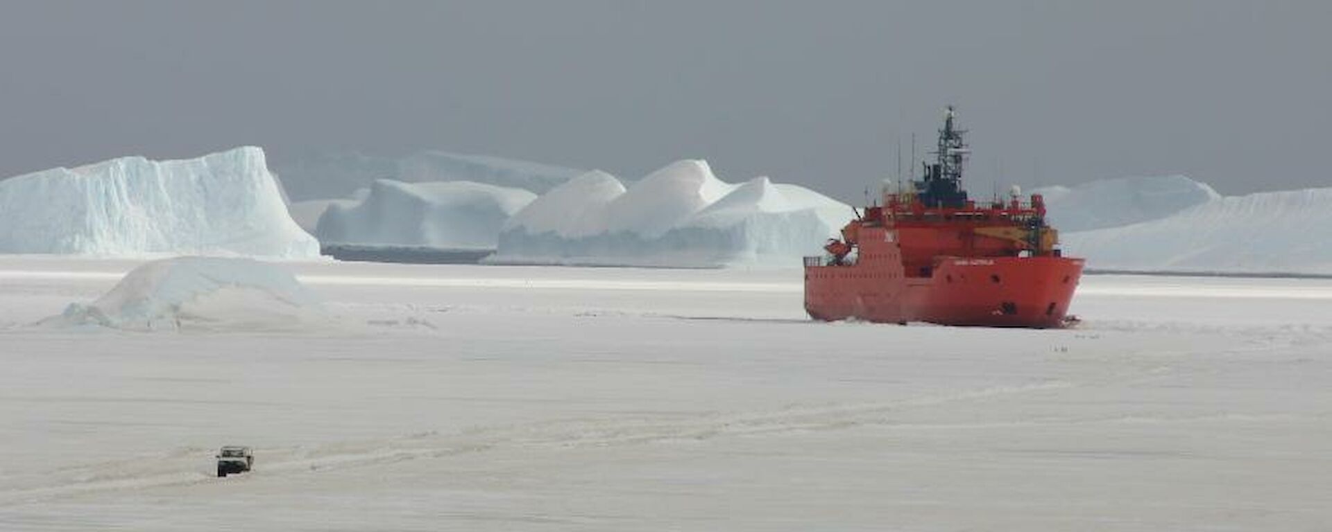 The brightly painted Aurora Australis parked up on the sea ice in front of the station with a vehicle travailing on the ice in the foreground