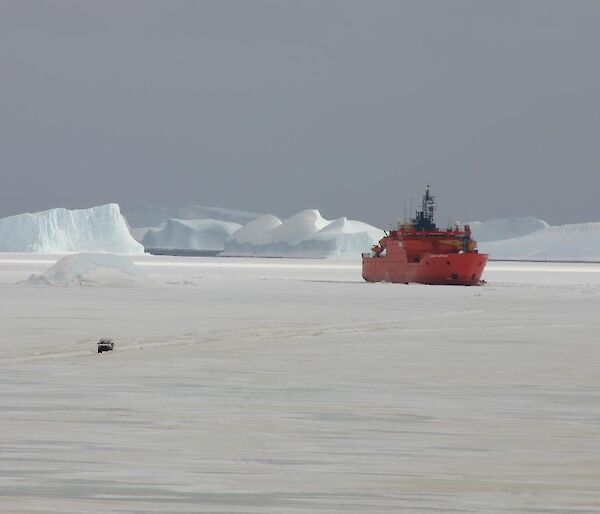 The brightly painted Aurora Australis parked up on the sea ice in front of the station with a vehicle travailing on the ice in the foreground