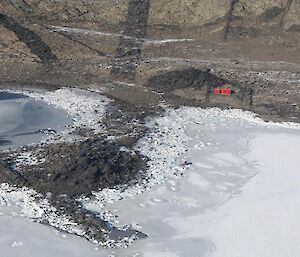 An aerial photo of the bright red Books hut standing out on the rocky terrain and the melting Fjord sea ice